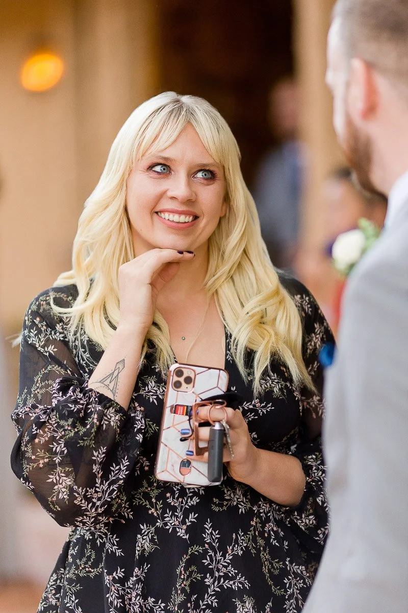 A woman with long blonde hair smiles warmly, holding a smartphone. She wears a black dress with a floral pattern, conveying joy in a warm setting.