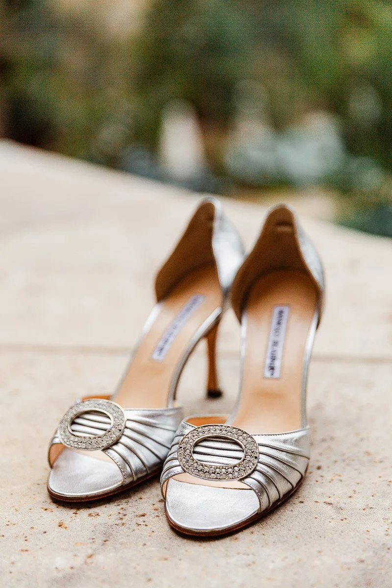 Silver high-heeled shoes with embellished round buckles, placed on a stone surface. Background blurred, conveying elegance and sophistication.