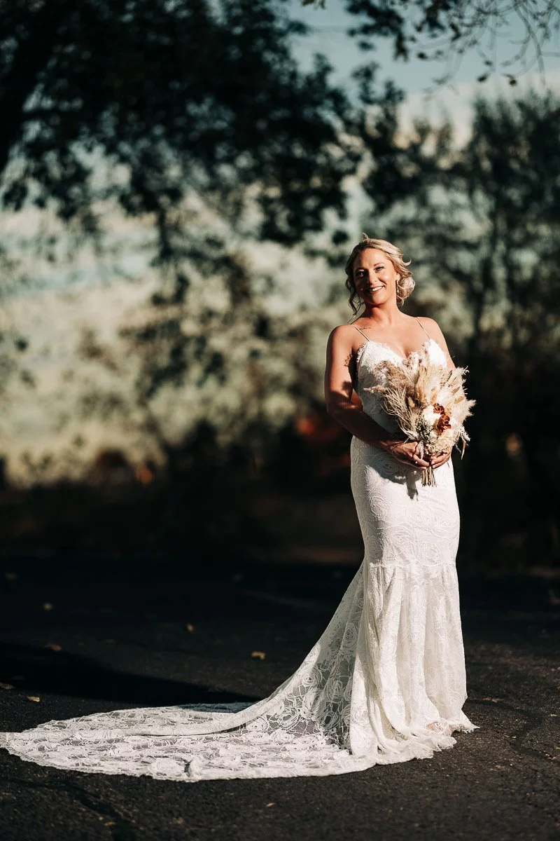 A bride stands outdoors in a lace wedding gown with a long train, holding a bouquet of dried flowers. She smiles under soft, natural lighting, evoking serenity.