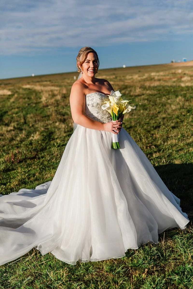 Bride in white gown holds yellow calla lilies, standing in a sunlit grassy field. Blue sky and horizon in the background, exuding joy and serenity.