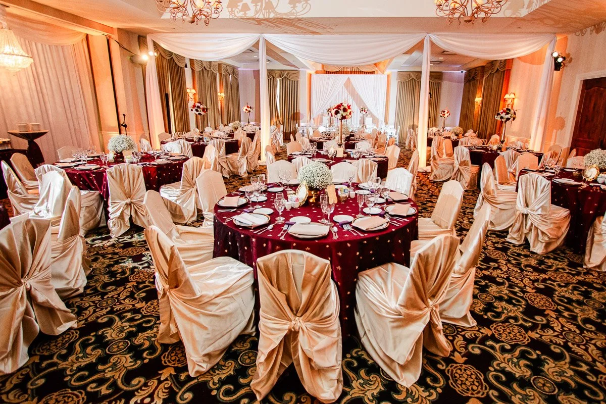 Elegant wedding reception hall with round tables, maroon tablecloths, white chair covers, floral centerpieces, and soft lighting creating a warm ambiance.