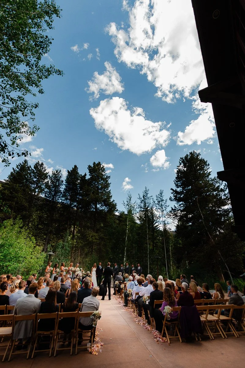 An outdoor wedding ceremony with guests seated on wooden chairs. A couple stands under a clear blue sky with scattered clouds, surrounded by lush green trees. The atmosphere is serene and romantic.