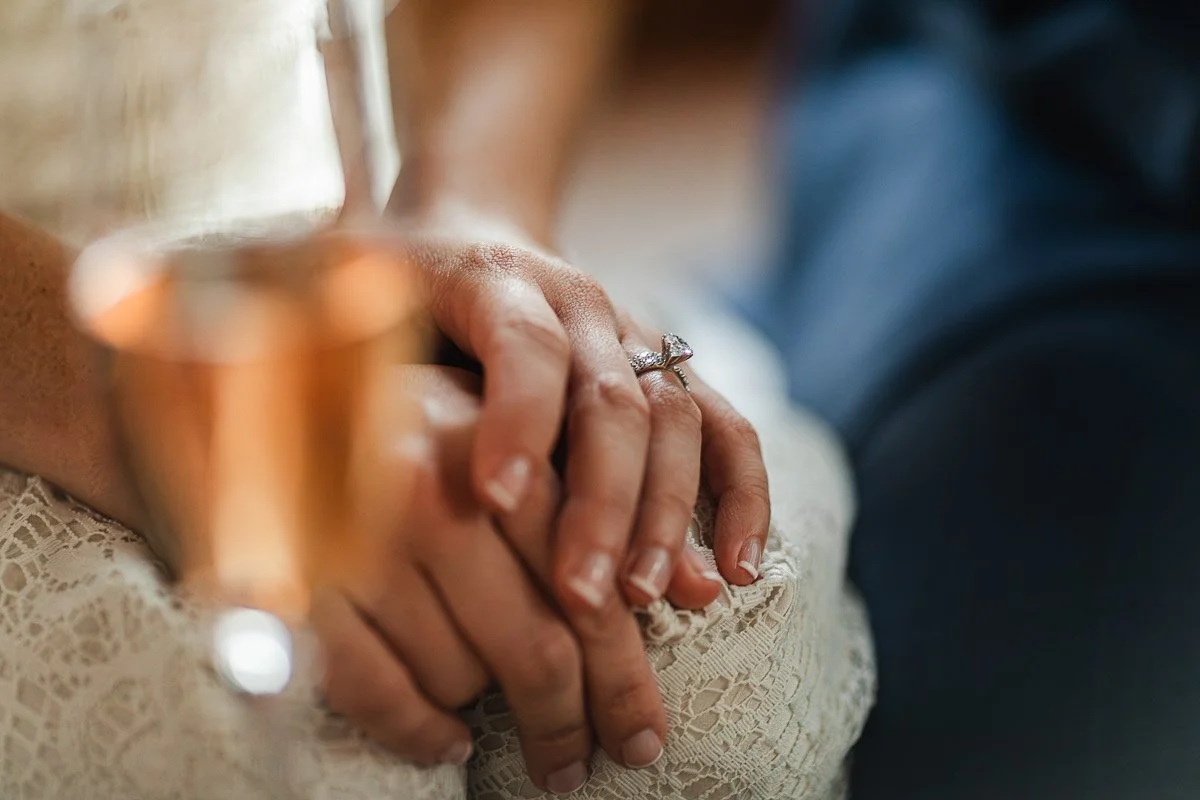 Close-up of two hands gently clasped, one with a diamond ring, resting on a lace fabric. A blurred champagne glass in the foreground suggests celebration.