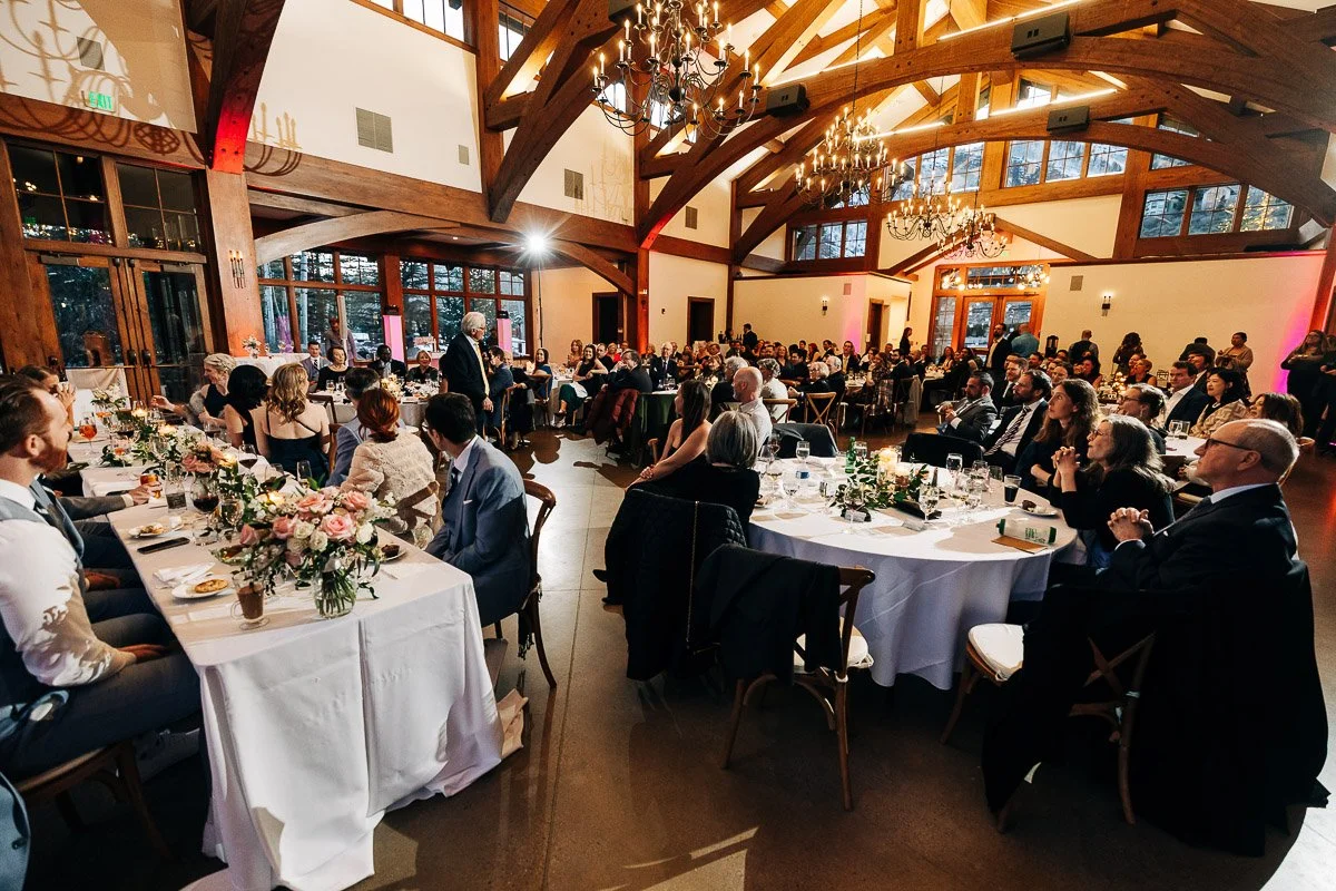 A wedding reception in a rustic hall with wooden beams and chandeliers. Guests seated at long, flower-adorned tables listen to a speaker.