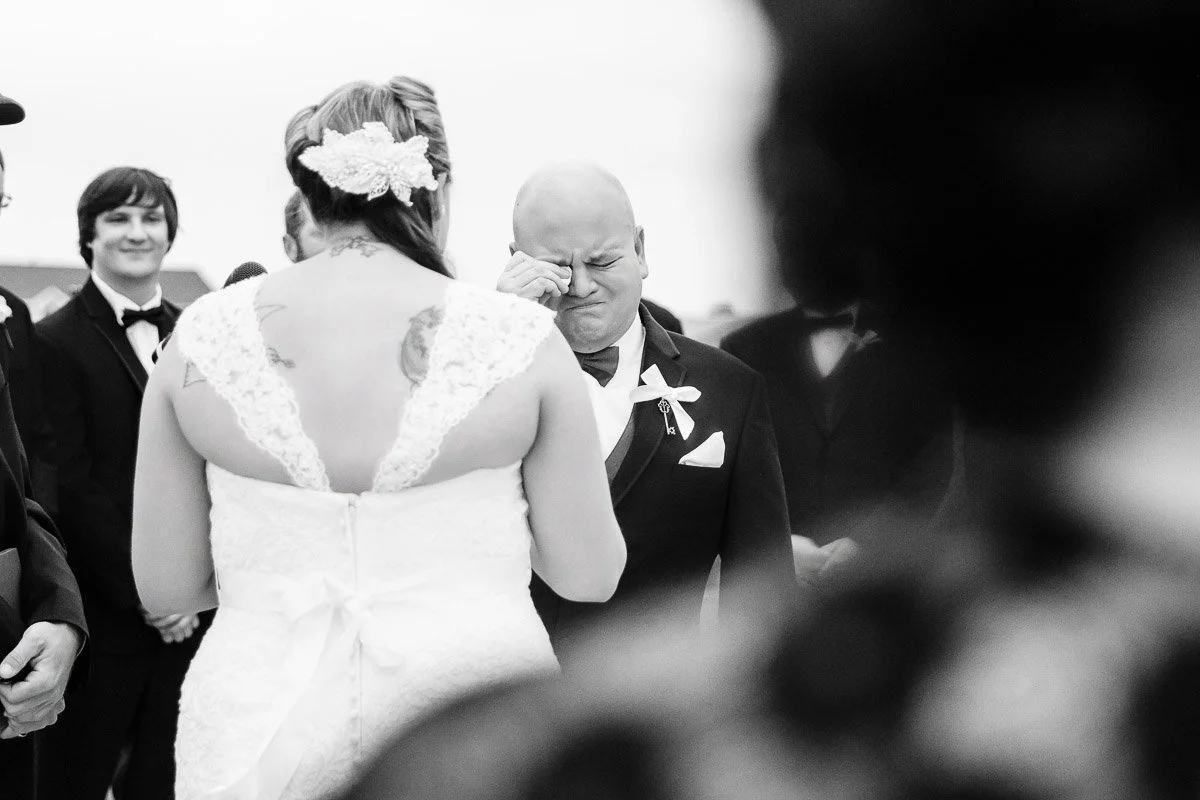 A groom wipes tears during an emotional wedding ceremony. The bride, seen from behind in a lace dress, stands facing him. Guests smile in the background.