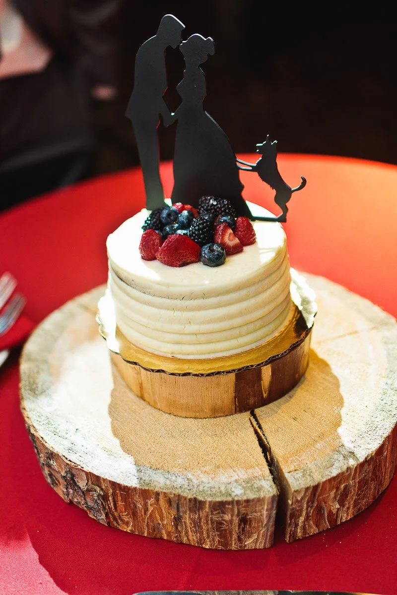 A round wedding cake with white frosting sits on a wooden slab. Topped with black silhouette figures of a couple and dog, adorned with fresh berries. Romantic tone.
