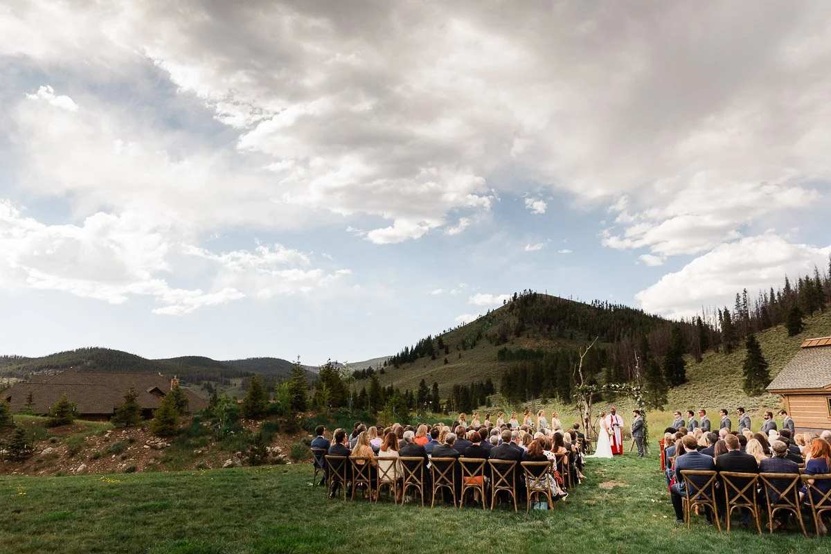 Outdoor wedding ceremony with guests seated on wooden chairs overlooking scenic mountains under a cloudy sky. The atmosphere is serene and joyful.