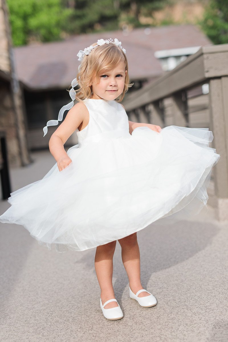 A young girl twirls in a white dress with a tulle skirt, wearing white shoes and a floral headband. She stands on an outdoor wooden deck, with soft sunlight and a blurred house in the background, conveying a joyful and playful mood.