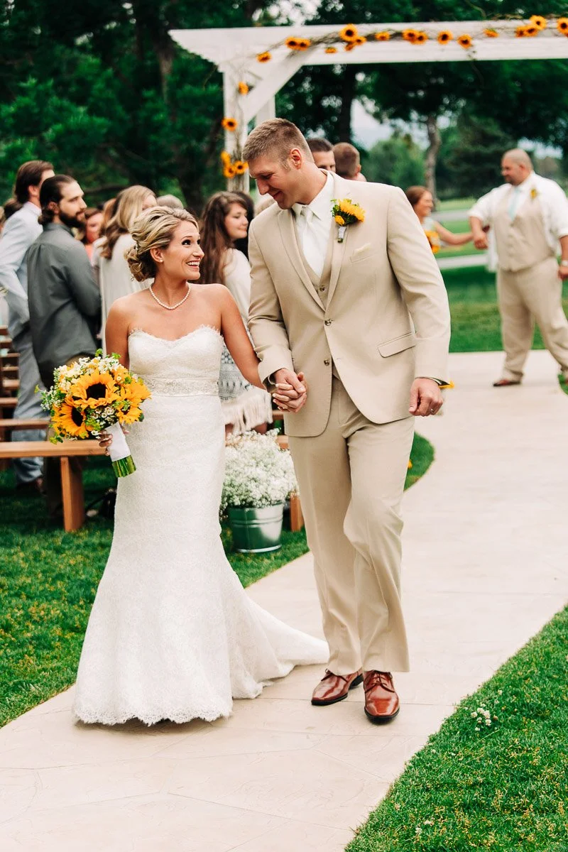 A joyful couple in wedding attire walks down an outdoor aisle, holding hands. The bride carries a sunflower bouquet, and guests watch them happily.