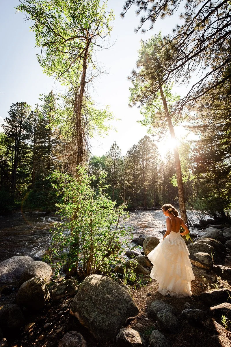 Bride in white gown stands by a forest stream, surrounded by tall trees and rocks, with sunlight streaming through the leaves. Serene and enchanting scene captured by Colorado wedding photographer tomKphoto