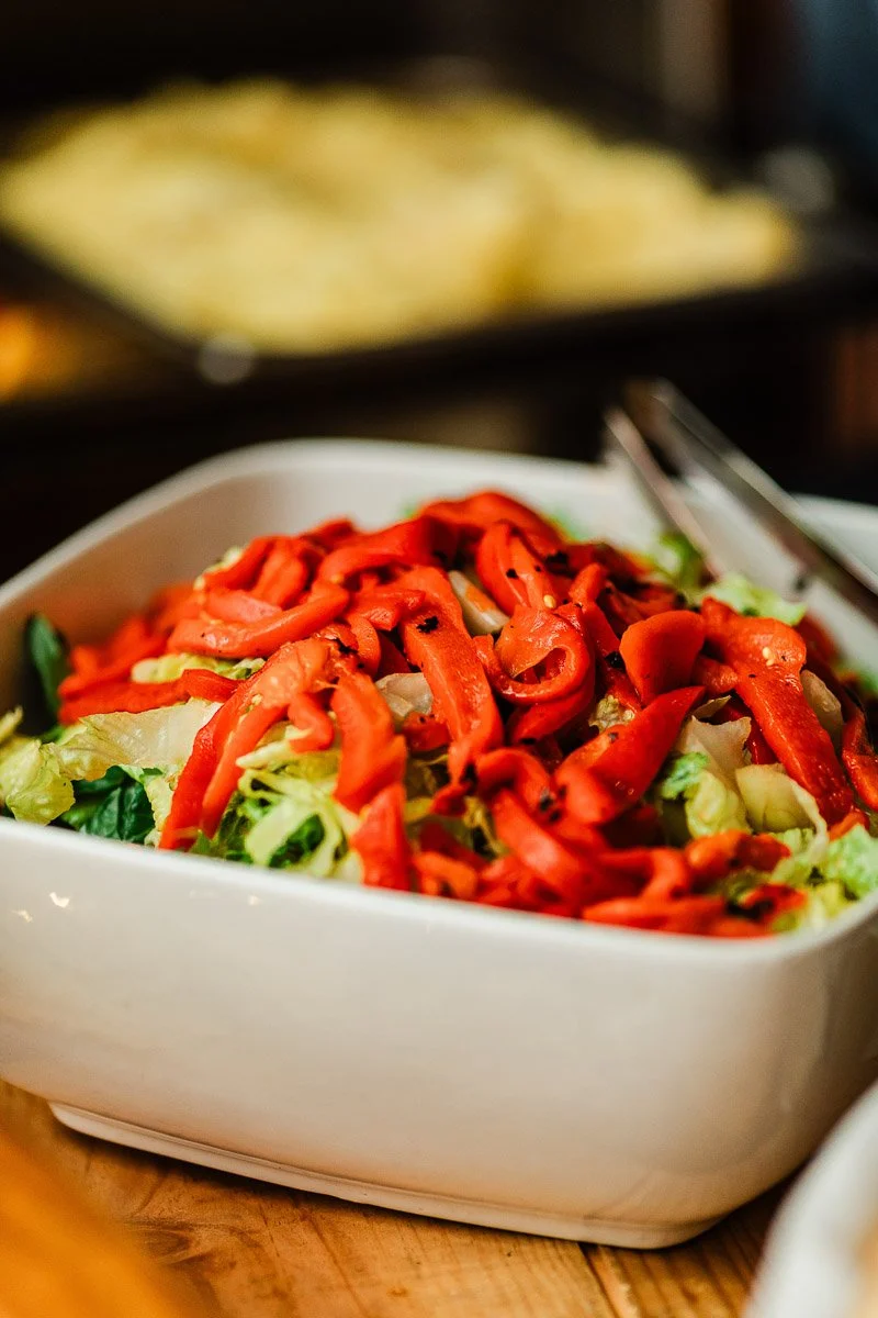 A white bowl filled with a fresh salad, featuring crisp lettuce and vibrant red pepper strips. Metal tongs rest on top, suggesting a ready-to-serve feel.