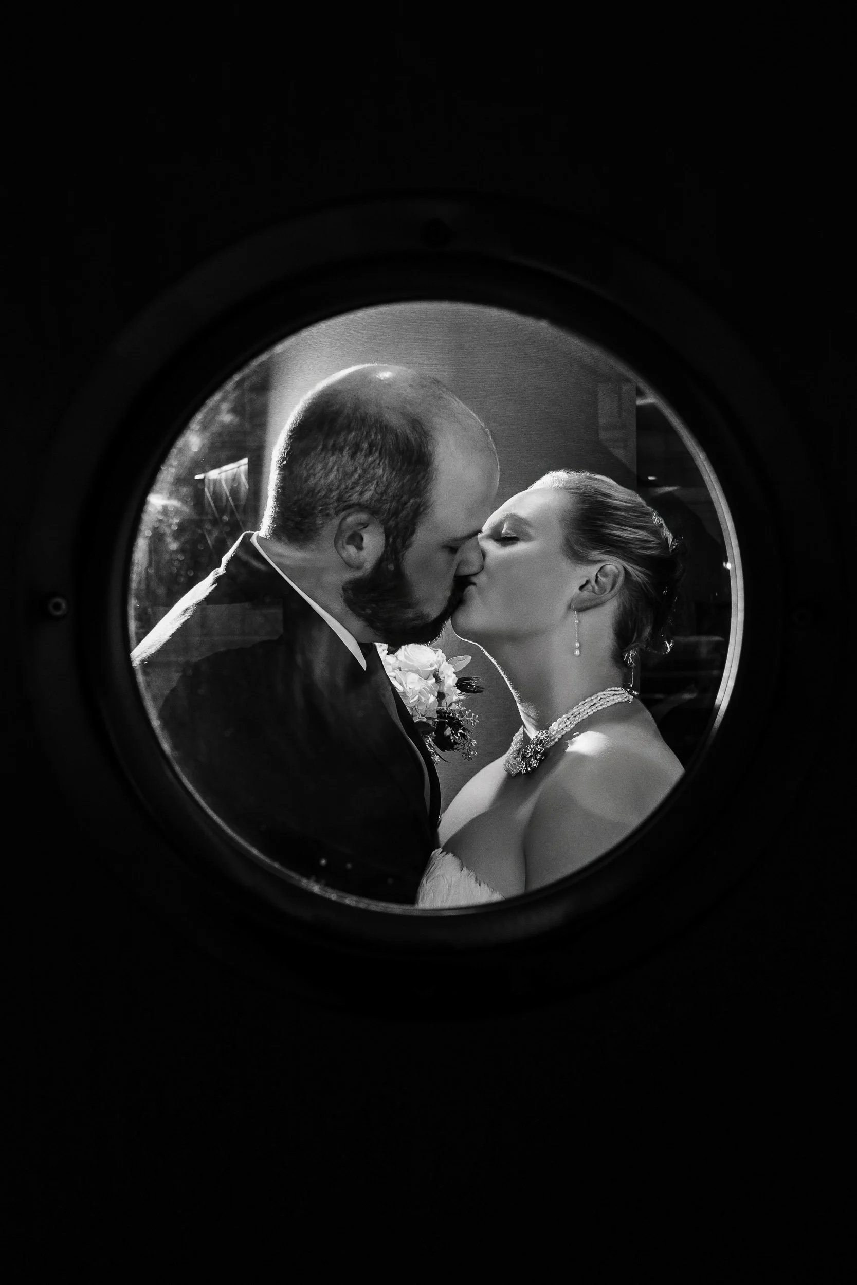 Bride and groom kiss in a porthole window during a Magnolia Hotel wedding in Denver, Colorado