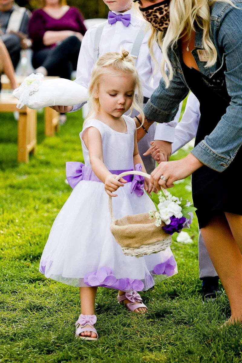 A young flower girl in a white dress with purple accents holds a basket. An adult helps her while a boy holds a cushion. The scene is cheerful and outdoors.