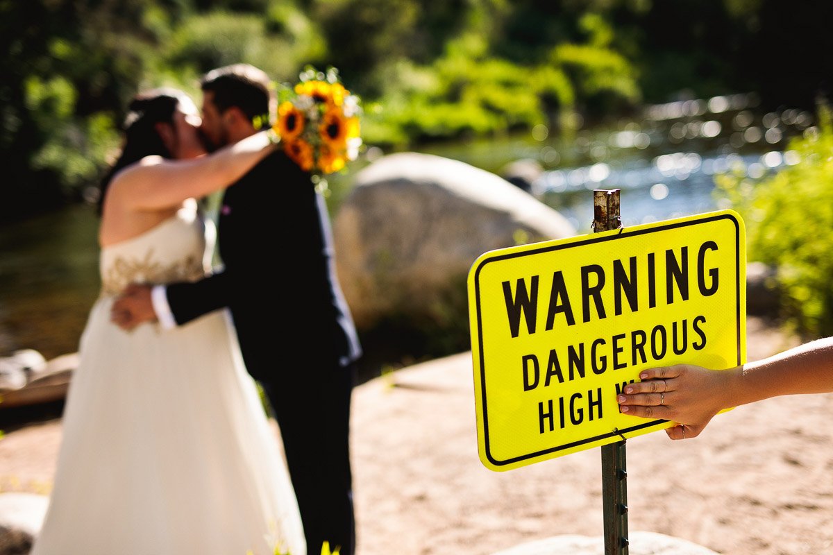 A couple kisses in wedding attire near a serene river. A bright yellow sign reading "Warning Dangerous High" adds humor to the romantic scene.