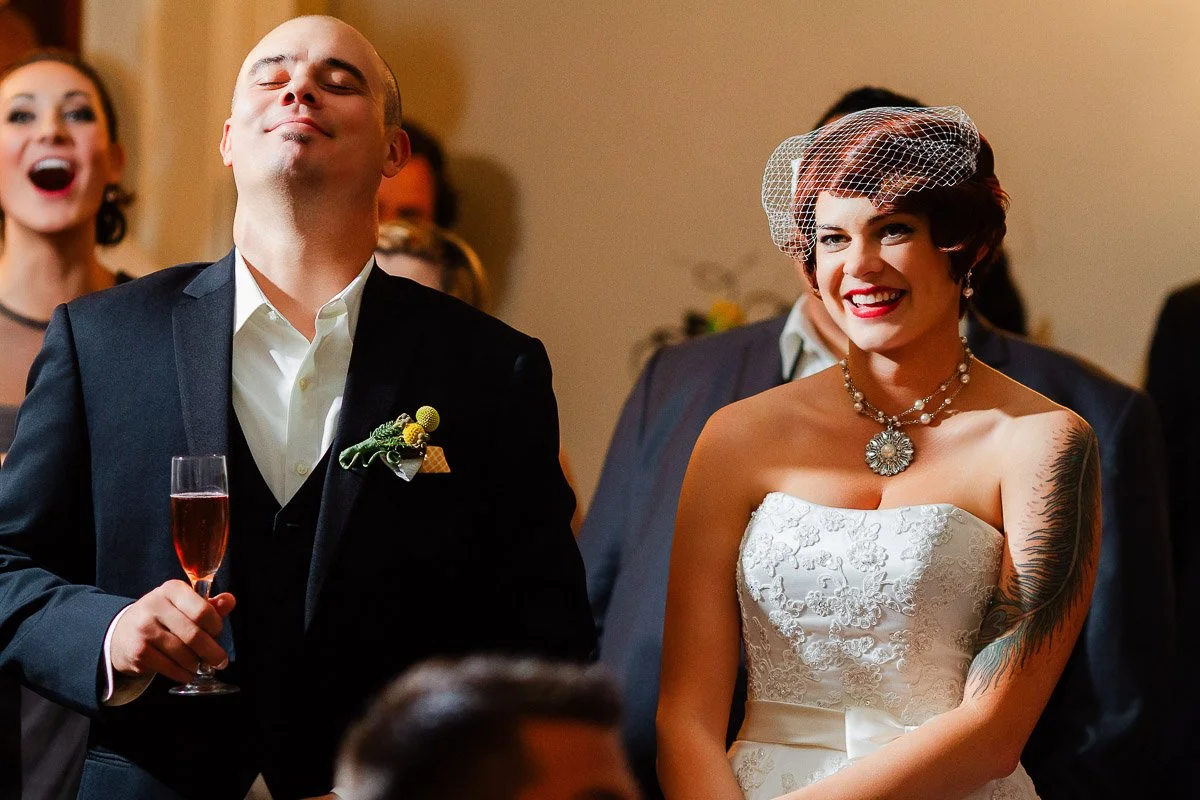A smiling bride in a white gown and birdcage veil stands beside a man in a suit holding a glass of champagne. The scene conveys joy and celebration.