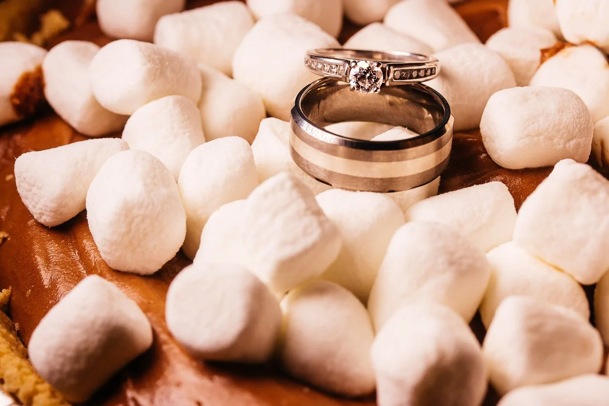 Two silver rings, one with a diamond, rest atop fluffy white marshmallows on a chocolate surface, evoking a sense of romance and sweetness.