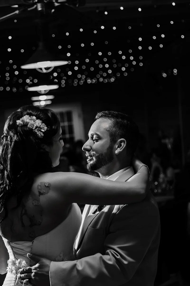 A couple shares a joyful dance at a wedding, embraced under twinkling fairy lights. The scene is intimate and warm, with soft smiles exchanged.