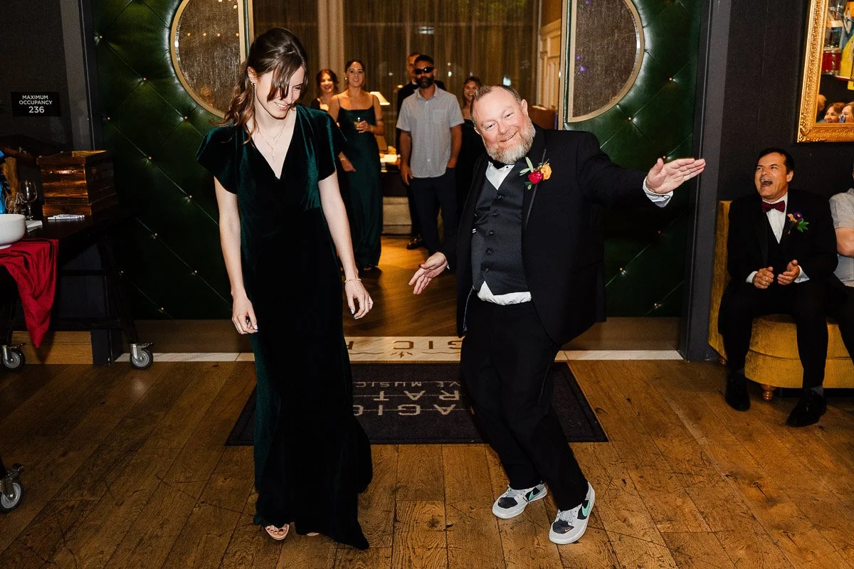 A man in a suit and sneakers dances joyfully beside a woman in a dark green dress at an indoor event. Onlookers smile in the background, creating a lively atmosphere.