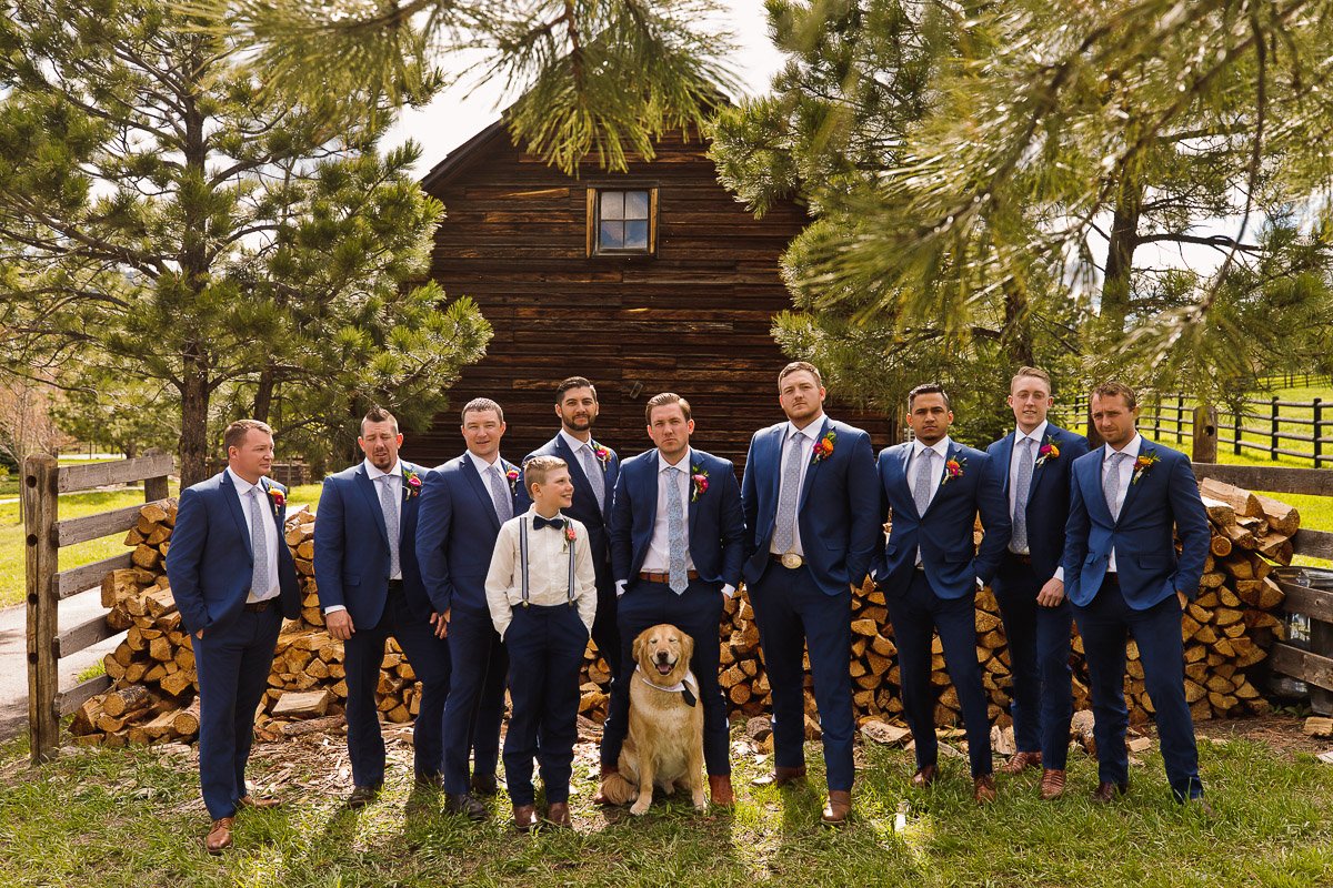 A group of groomsmen in blue suits and a boy in suspenders pose with a dog in front of a rustic cabin, surrounded by greenery and logs at a Spruce Mountain Ranch wedding in Larkspur, Colorado