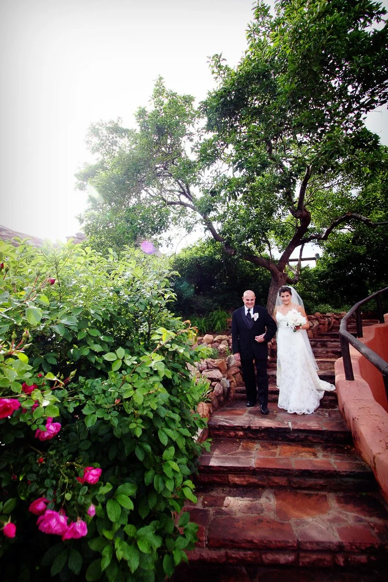 A bride in a white lace gown and veil walks arm-in-arm with a man in a tuxedo down stone steps. They are surrounded by lush greenery and pink flowers, creating a serene, joyful mood captured by Red Rocks Wedding Photographer tomKphoto