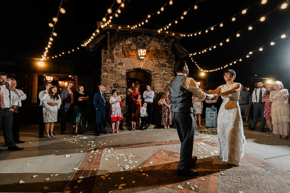 A bride and groom share their first dance outdoors at night, under warm string lights. Guests watch in admiration, creating a joyful, romantic atmosphere.