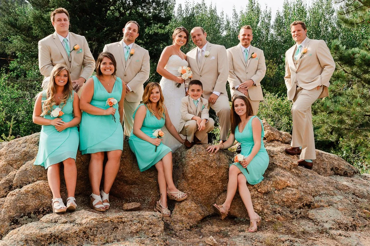 A wedding party poses joyfully on a rocky terrain. The bride in white and bridesmaids in turquoise dresses hold bouquets; groomsmen wear beige suits. Lush greenery surrounds them.