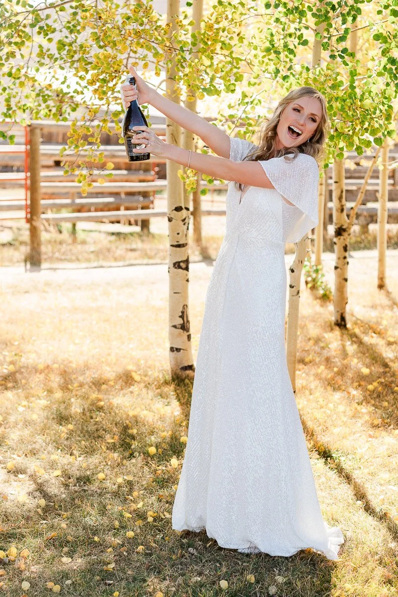 Bride in a white gown joyfully holding a champagne bottle among sunlit autumn trees, exuding happiness and celebration in a serene outdoor setting.