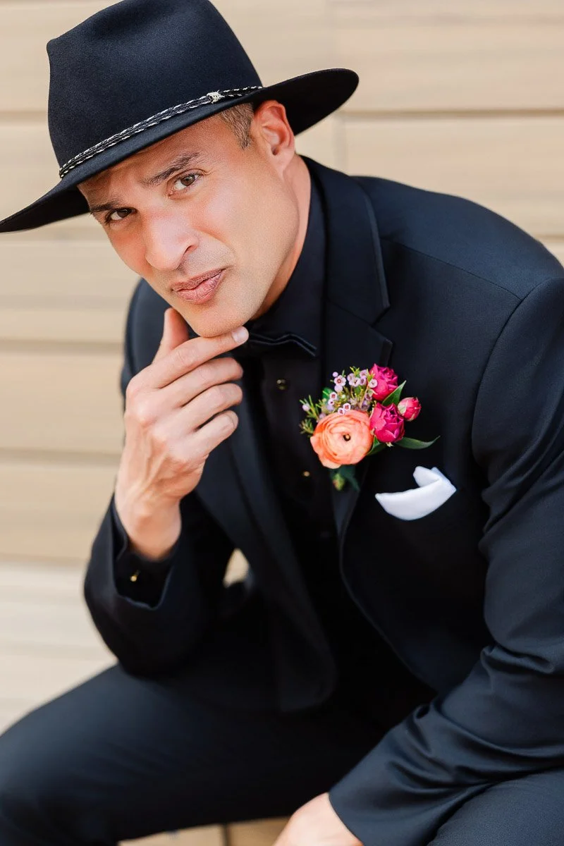 A man in a black suit and cowboy hat sits, hand on chin, exuding confidence. He wears a vibrant flower boutonniere, contrasting with the neutral backdrop.