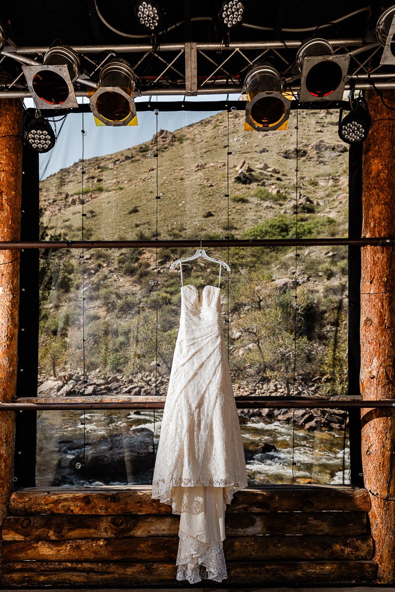 A lace wedding dress hangs on the stage of the Mishawalka Amphitheatre with a scenic view of a hillside and flowing river, creating a serene and romantic atmosphere.