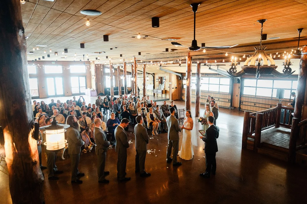 Wedding ceremony in a rustic hall with wood beams and chandeliers. Bride and groom at the altar, surrounded by guests seated in rows. Warm, festive atmosphere at a Spruce Mountain Ranch wedding in Larkspur, Colorado