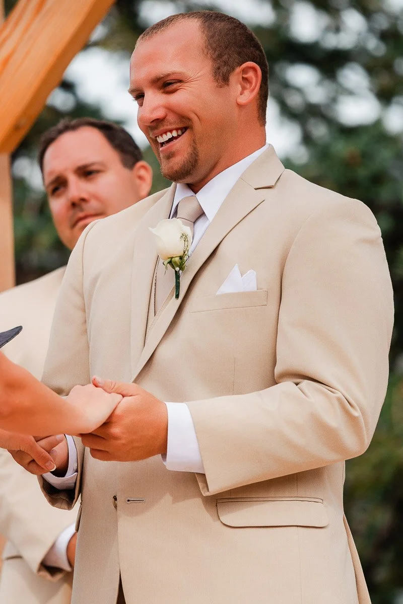 A man in a beige suit, smiling warmly, holds hands with someone at a wedding. He wears a white rose boutonniere. Another man observes in the background.