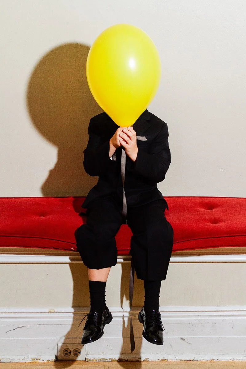 A child in a black suit sits on a red bench, holding a large yellow balloon covering their face. The setting is minimal, with bright lighting.