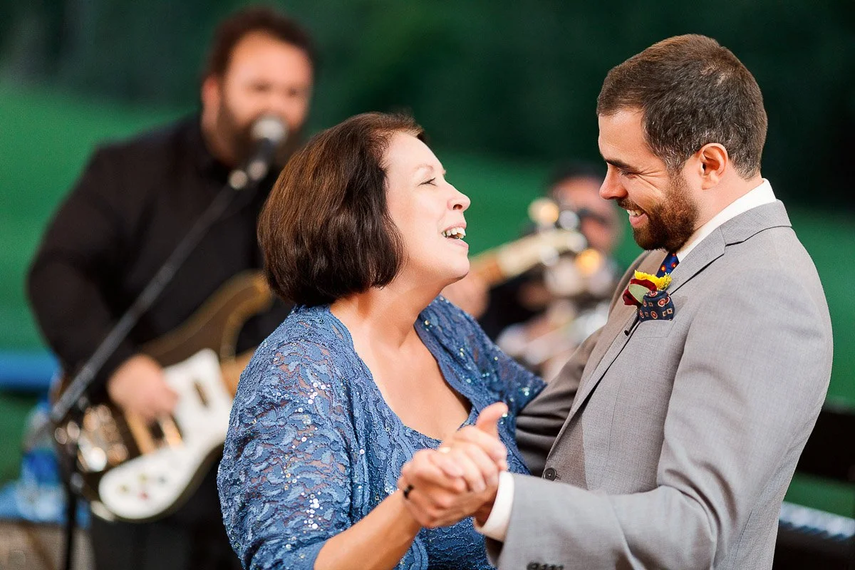 A woman in a blue dress and a man in a gray suit joyfully dance at an outdoor event, smiling warmly at each other. A guitarist plays in the background.