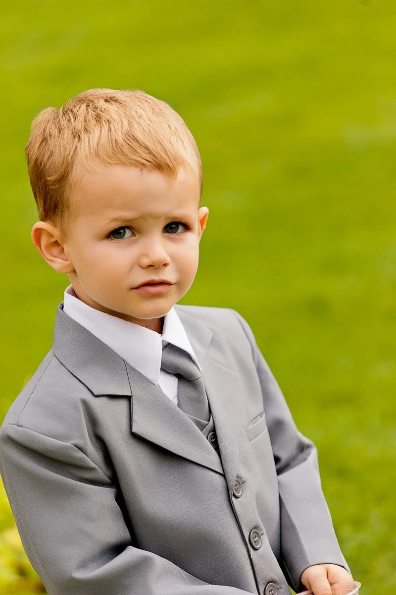 A young boy with light brown hair wears a gray suit, white shirt, and tie, standing on a green lawn. He looks curious and slightly serious.