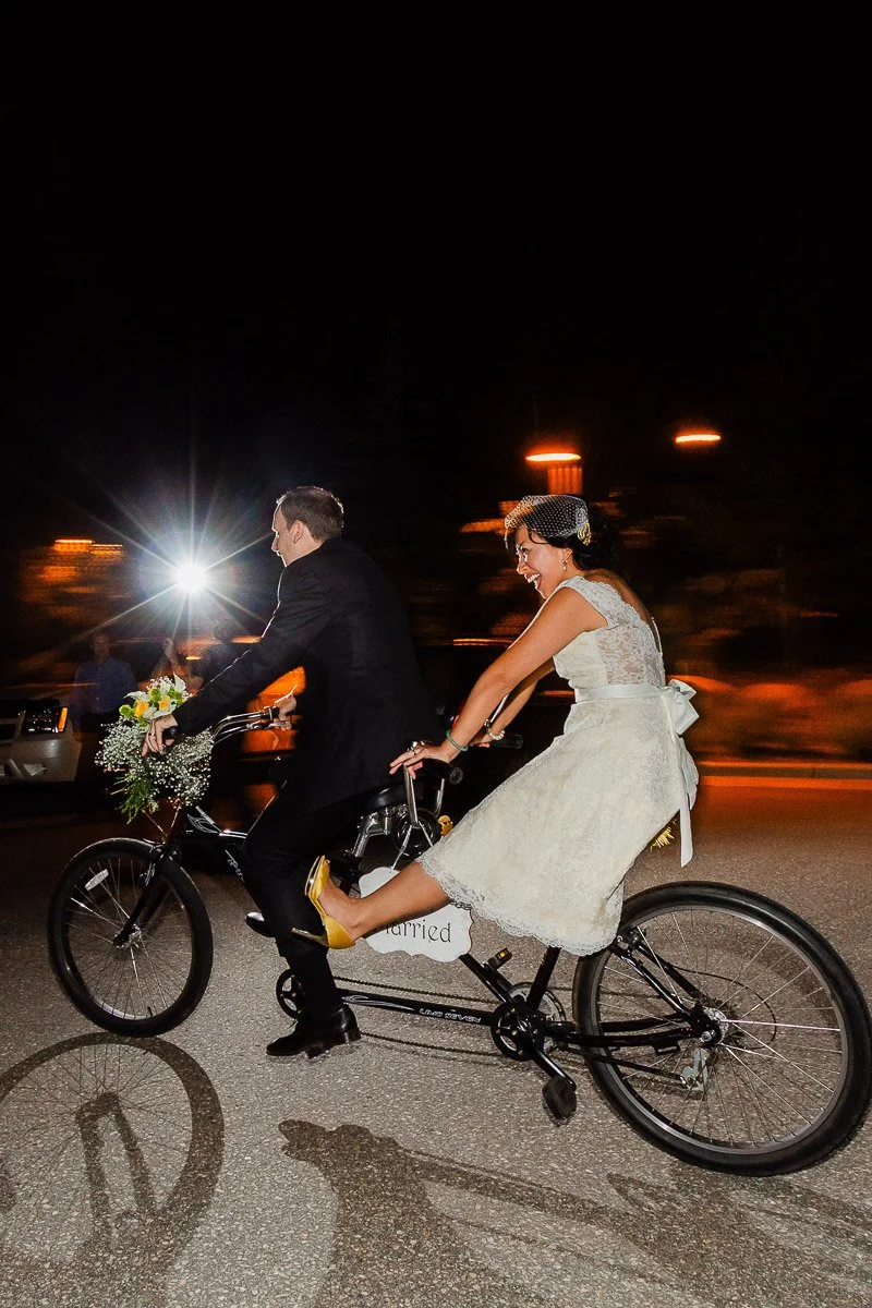 Bride and groom on tandem bike at night; she in a white dress, laughing with joy, a bouquet on handlebars. A flash captures their wedding exit concluding a Donovan Pavilion wedding in Vail, Colorado