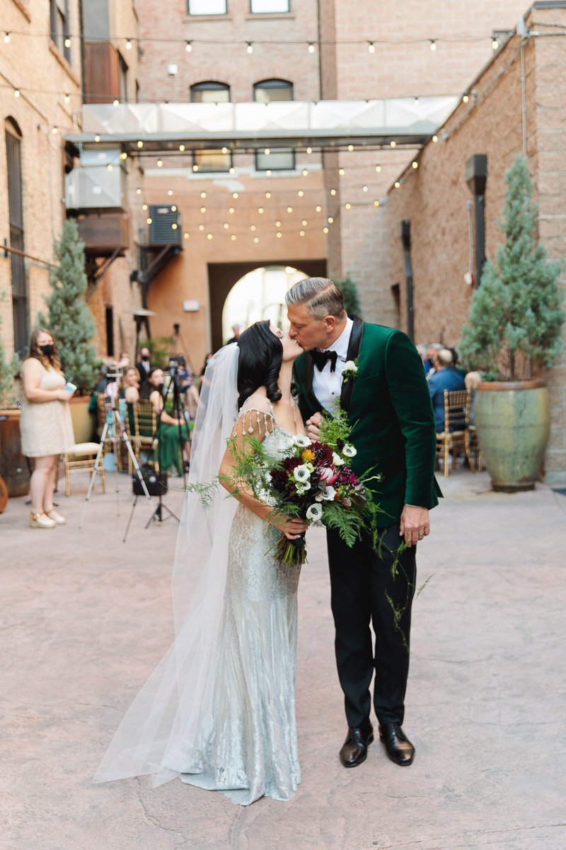 Bride in a shimmering gown and groom in a green suit share a kiss in a charming outdoor venue. Soft lights and guests create a romantic atmosphere.