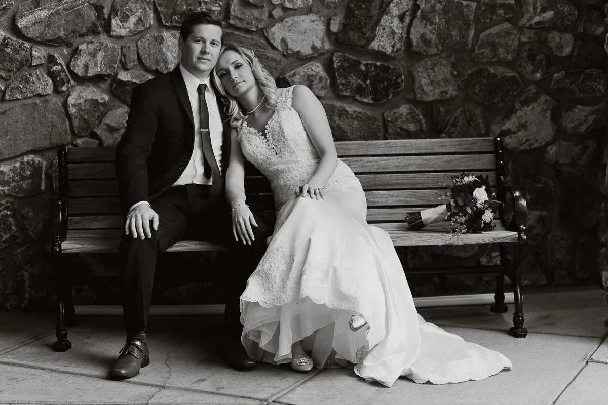 A couple in wedding attire sits on a bench against a stone wall. The bride leans on the groom's shoulder, holding his hand, next to a bouquet.