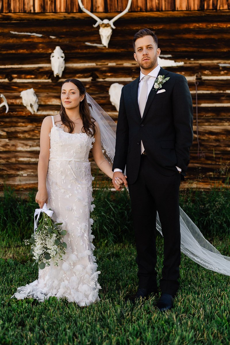 A bride and groom stand on grass in front of a rustic wooden wall adorned with animal skulls. The bride holds a bouquet, her dress detailed and white, while the groom wears a black suit, both with neutral expressions.