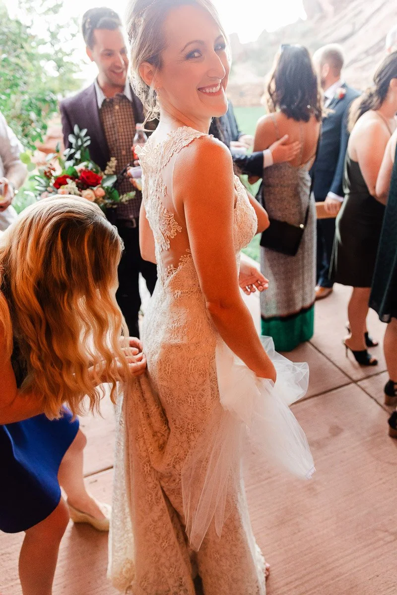 Bride in lace gown smiling, surrounded by guests at a wedding. A woman adjusts her dress. The scene is joyful, with a lively outdoor atmosphere.