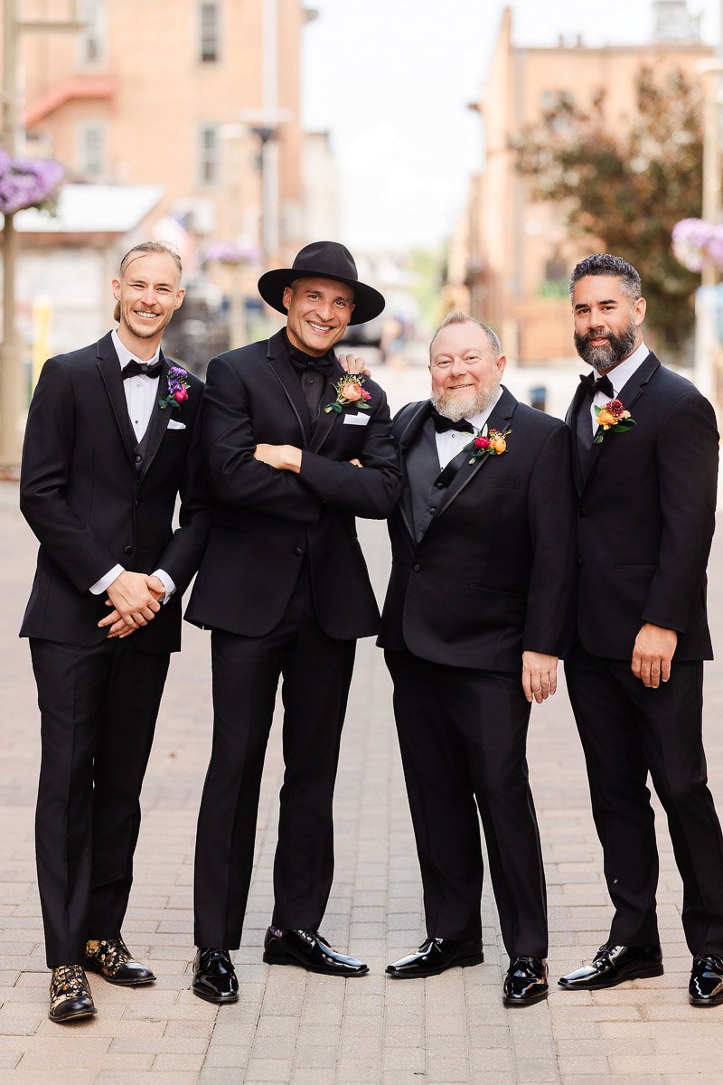Four men in black tuxedos stand closely on a charming cobblestone street. They are smiling warmly, conveying a joyful and celebratory mood.