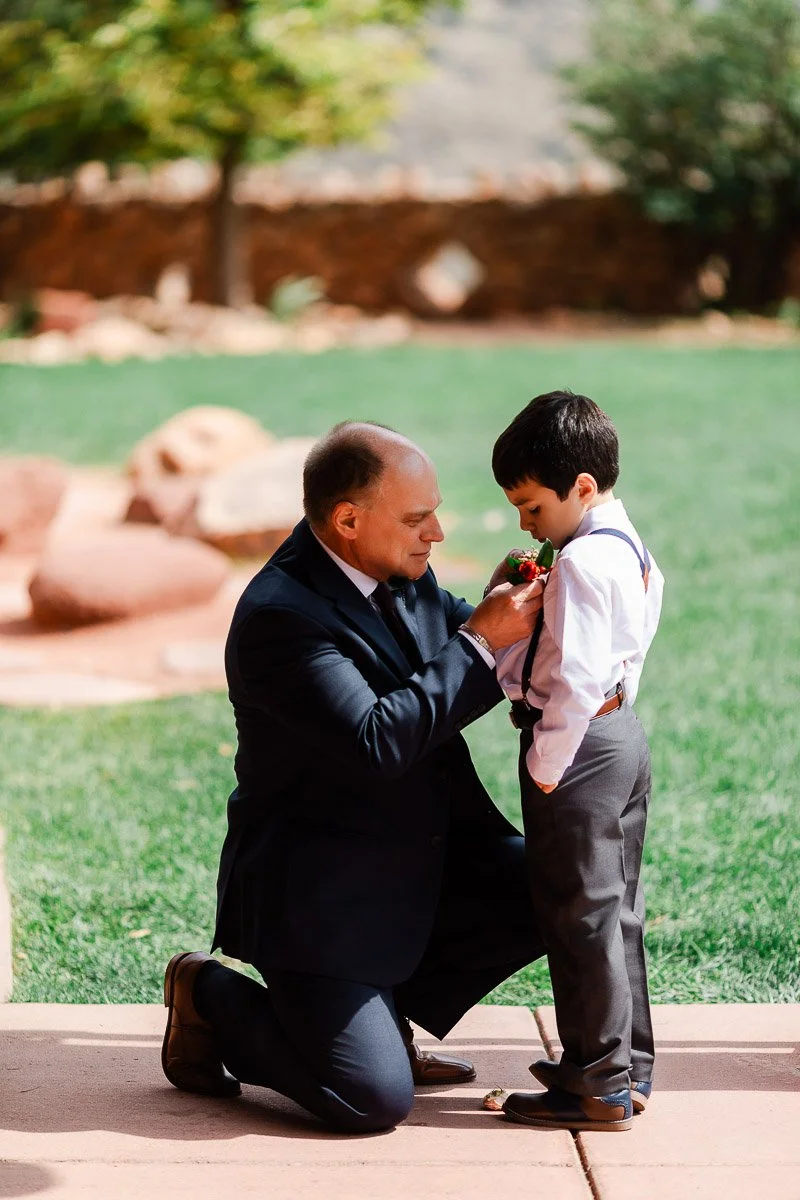 A man kneels on a patio, helping a young boy adjust a red boutonniere on his shirt. They share a tender moment in a garden setting.