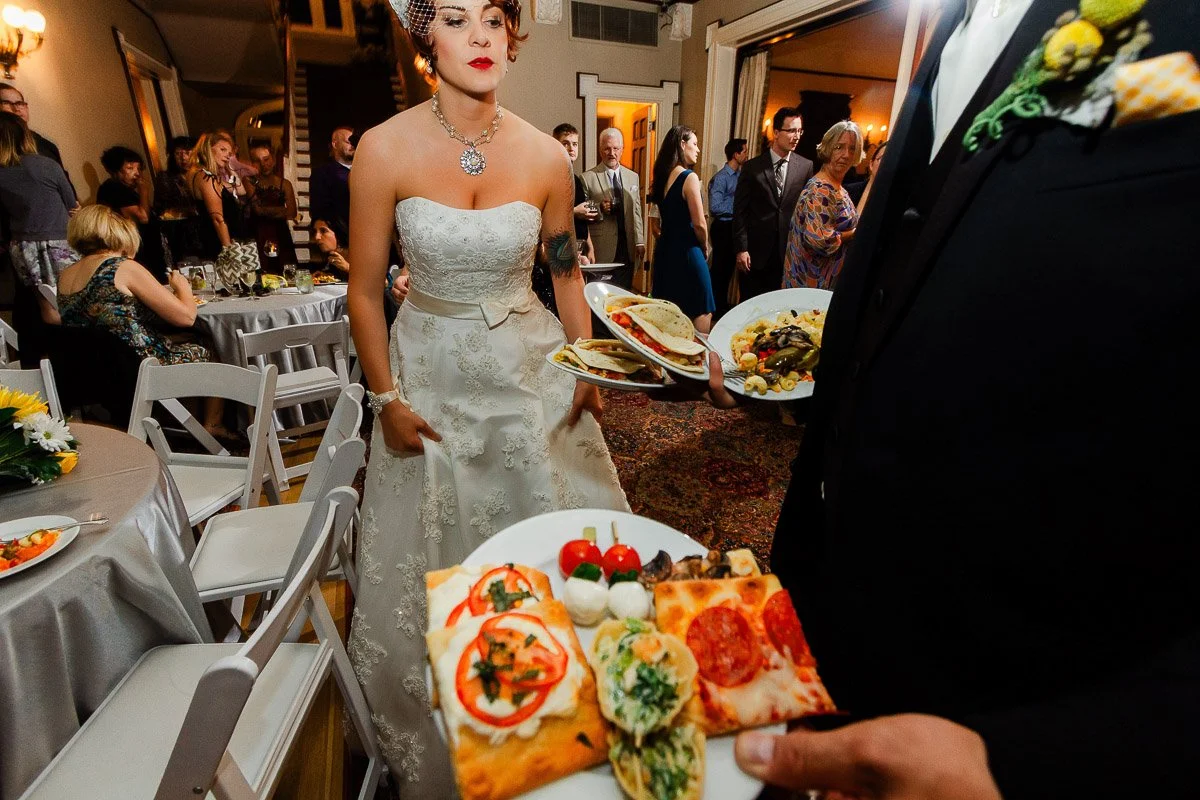 A bride in a white dress stands at a lively wedding reception, facing someone holding plates of assorted food, including pizza and skewers. The atmosphere is festive.
