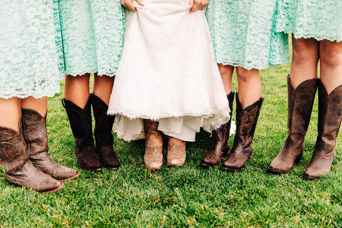 Bridesmaids in mint green lace dresses and cowboy boots stand on grass, surrounding a bride in a white gown and boots, evoking a rustic vibe at a Barn at Raccoon Creek wedding in Littleton, Colorado
