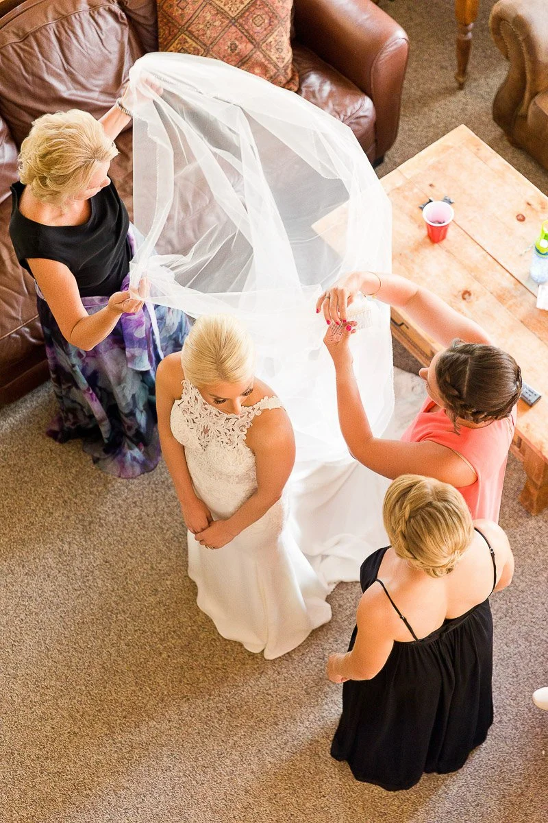 Bride in elegant lace gown surrounded by three women adjusting her long veil. Warm, joyful atmosphere in living room with cozy furniture.
