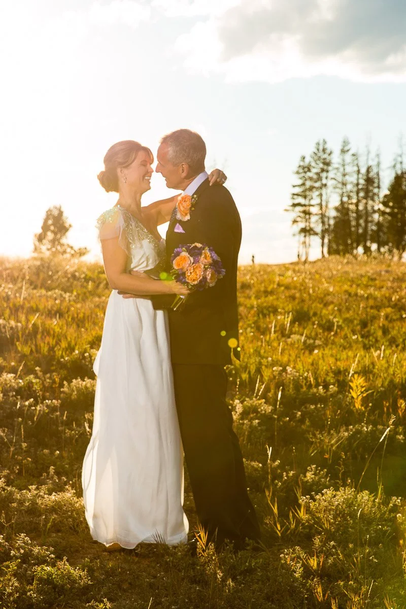 A couple in wedding attire stands in a sunlit field, embracing and smiling warmly. The bride holds a bouquet of colorful flowers. Trees and a bright sky are in the background, conveying a joyful, serene atmosphere.