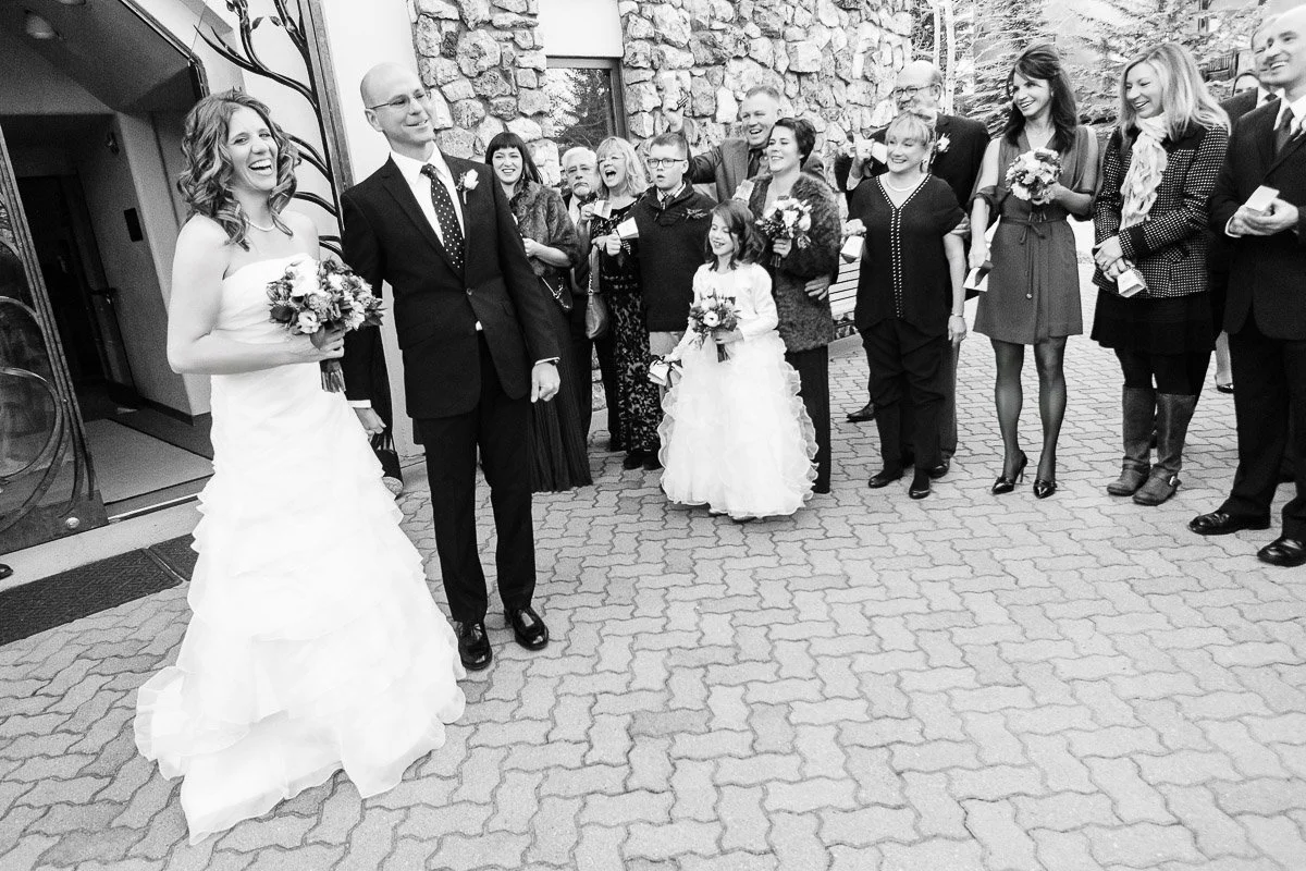 A joyful bride and groom stand in front of a cheering crowd outside. The bride holds a bouquet, and a flower girl stands nearby. Laughter and celebration.