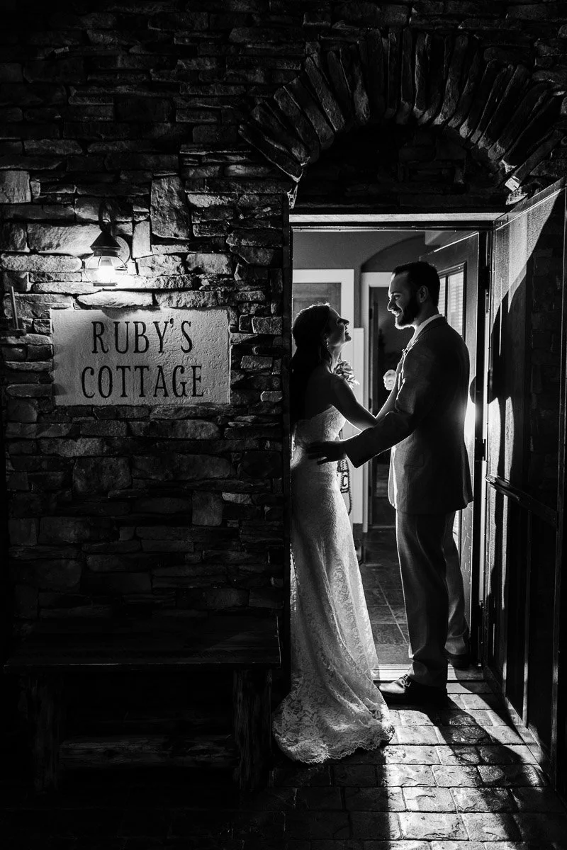 A couple stands in a doorway, embracing with tender expressions. The scene is lit dramatically from behind. A "Ruby's Cottage" sign is visible on stone walls at night during a Spruce Mountain Ranch wedding in Larkspur, Colorado