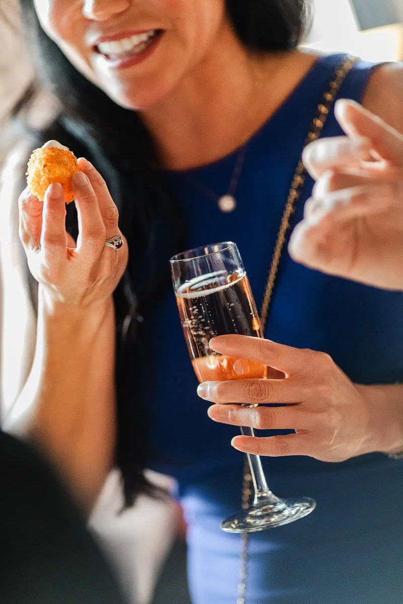 A woman in a blue dress holds a glass of rosé champagne and a small appetizer, smiling and engaged in conversation. The scene is lively and social.
