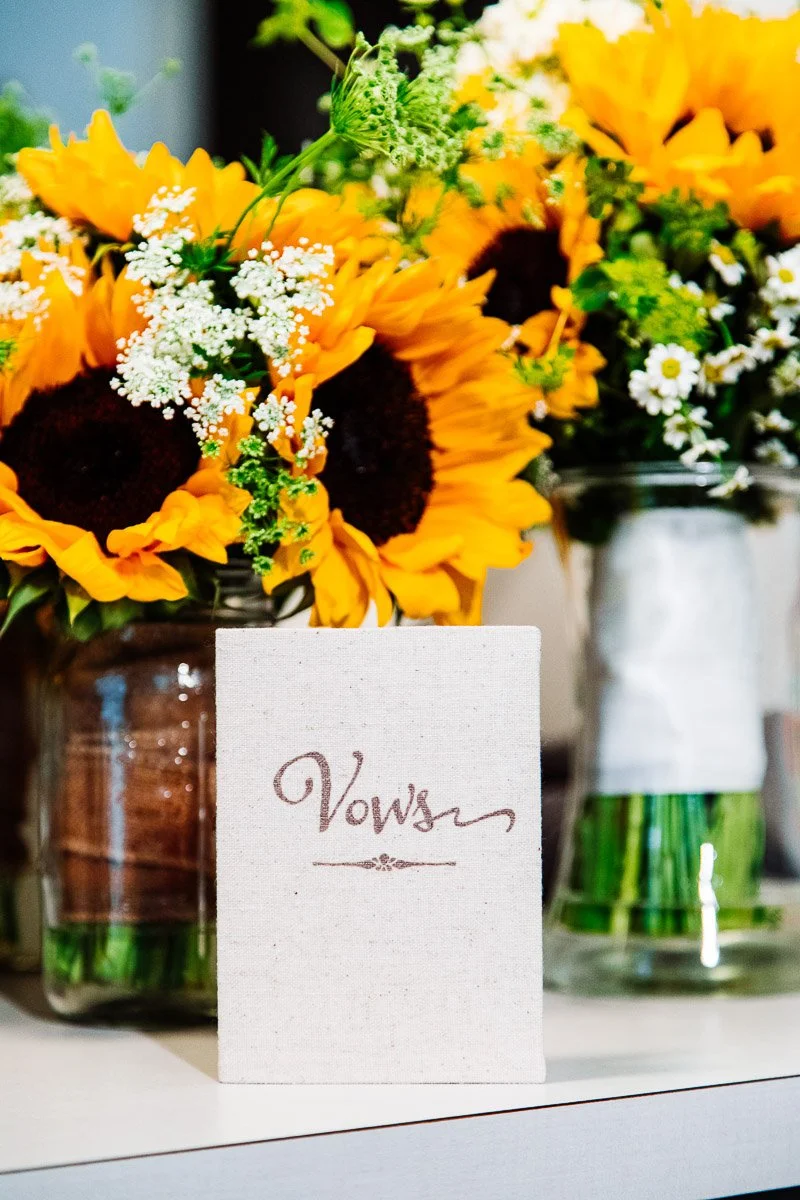 A small canvas book labeled "Vows" stands in front of vibrant sunflower bouquets in glass jars. The scene is warm and cheerful, evoking love and celebration.