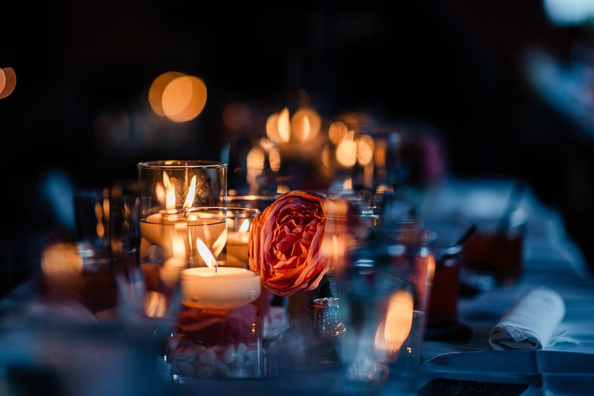 A soft, romantic table setting with glowing candles and a pink rose, creating a warm, intimate ambiance. The background is softly blurred.
